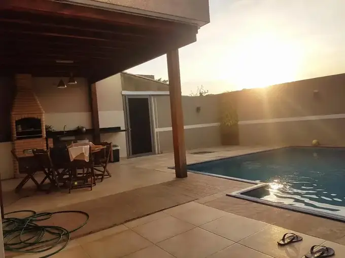 Área gourmet com churrasqueira, pergolado de madeira e piscina ao entardecer